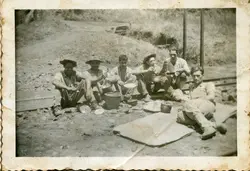 Uma fotografia em preto e branco que retrata um grupo de seis homens fazendo uma refeição ao ar livre, possivelmente trabalhadores ferroviários ou rurais em uma pausa. Eles estão sentados diretamente no solo, próximo a trilhos, utilizando utensílios simples como panelas e pratos, vestindo roupas de trabalho típicas da primeira metade do século XX.
