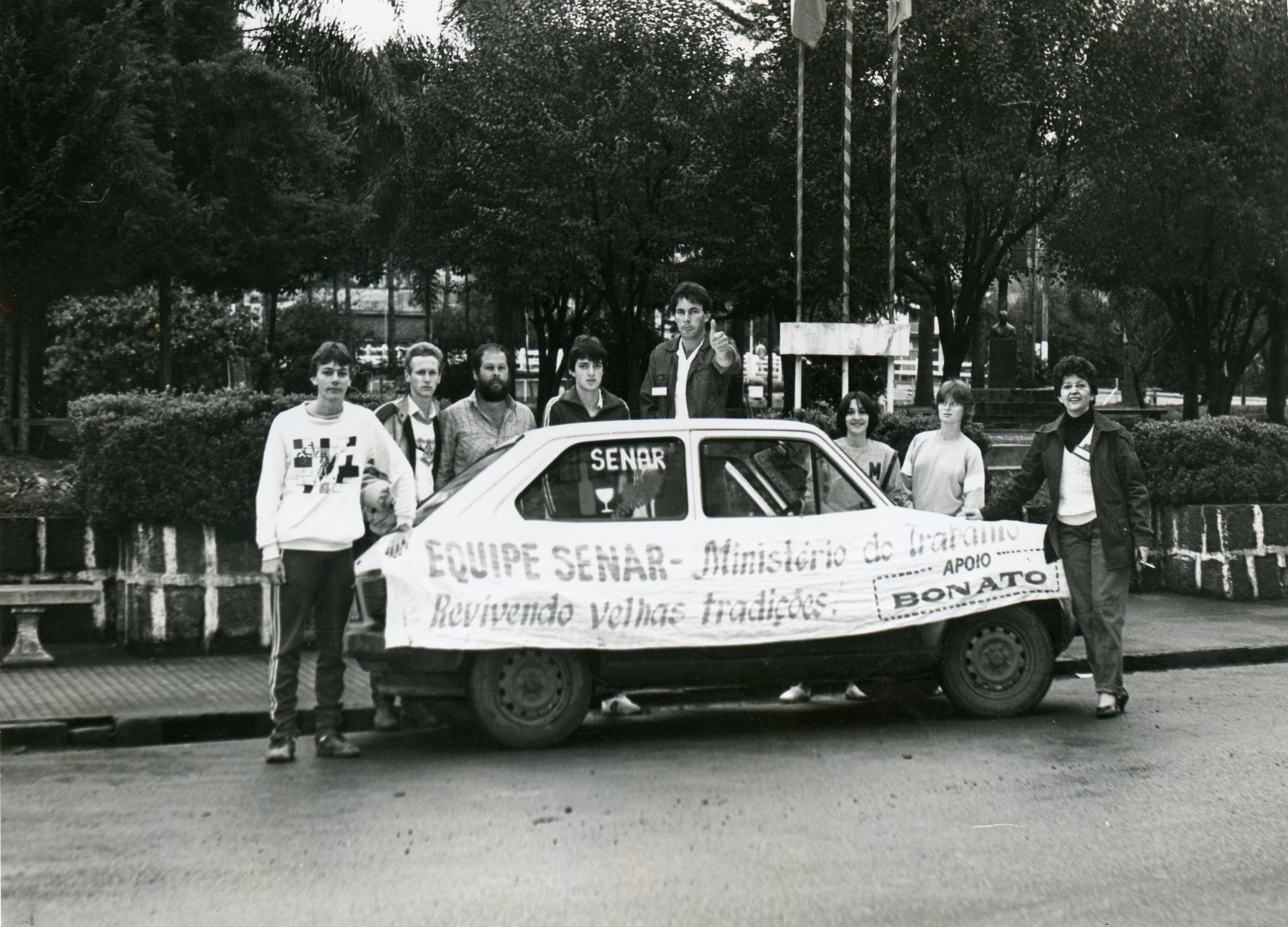Uma fotografia em preto e branco mostrando um grupo de pessoas posando ao lado de um automóvel compacto em uma praça ou área pública. O veículo exibe uma faixa com os dizeres 'Equipe SENAR - Ministério do Trabalho' e 'Revivendo velhas tradições', sugerindo uma ação comunitária ou evento educativo provavelmente ocorrido entre as décadas de 1980 e 1990.