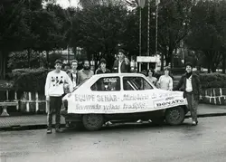 Uma fotografia em preto e branco mostrando um grupo de pessoas posando ao lado de um automóvel compacto em uma praça ou área pública. O veículo exibe uma faixa com os dizeres 'Equipe SENAR - Ministério do Trabalho' e 'Revivendo velhas tradições', sugerindo uma ação comunitária ou evento educativo provavelmente ocorrido entre as décadas de 1980 e 1990.