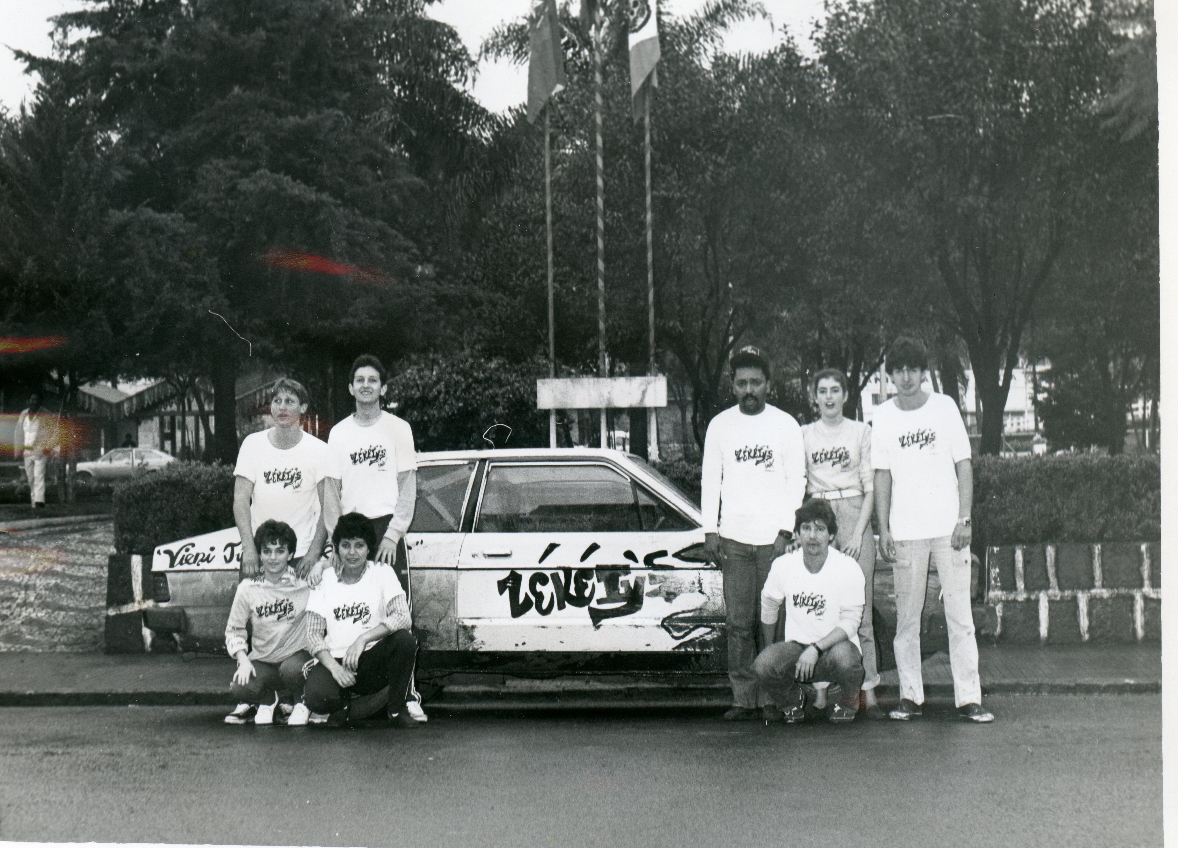 Uma fotografia em preto e branco mostrando um grupo de jovens posando ao lado de um carro personalizado com a inscrição 'Zekey's'. O grupo veste camisetas uniformes, sugerindo uma equipe ou evento promocional em um ambiente urbano, possivelmente uma praça ou área pública, com bandeiras ao fundo.