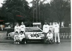 Uma fotografia em preto e branco mostrando um grupo de jovens posando ao lado de um carro personalizado com a inscrição 'Zekey's'. O grupo veste camisetas uniformes, sugerindo uma equipe ou evento promocional em um ambiente urbano, possivelmente uma praça ou área pública, com bandeiras ao fundo.