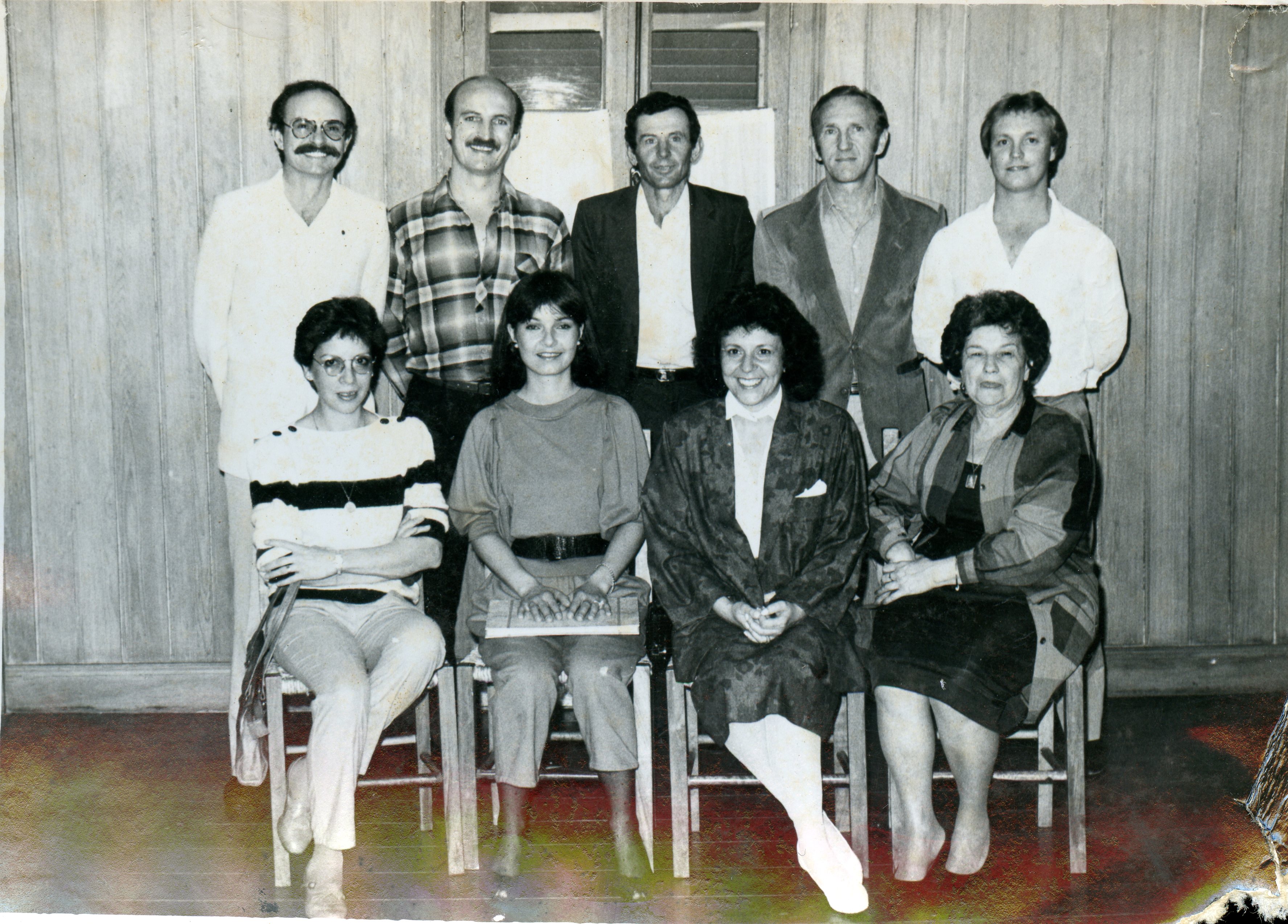 Uma fotografia de grupo composta por nove pessoas, quatro homens em pé e cinco mulheres sentadas, posando em um ambiente interno com paredes de madeira. O estilo das vestimentas, incluindo camisas de flanela, blazers e cortes de cabelo, sugere uma fotografia tirada durante a década de 1980 em um contexto social ou profissional brasileiro.