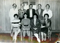 Uma fotografia de grupo composta por nove pessoas, quatro homens em pé e cinco mulheres sentadas, posando em um ambiente interno com paredes de madeira. O estilo das vestimentas, incluindo camisas de flanela, blazers e cortes de cabelo, sugere uma fotografia tirada durante a década de 1980 em um contexto social ou profissional brasileiro.