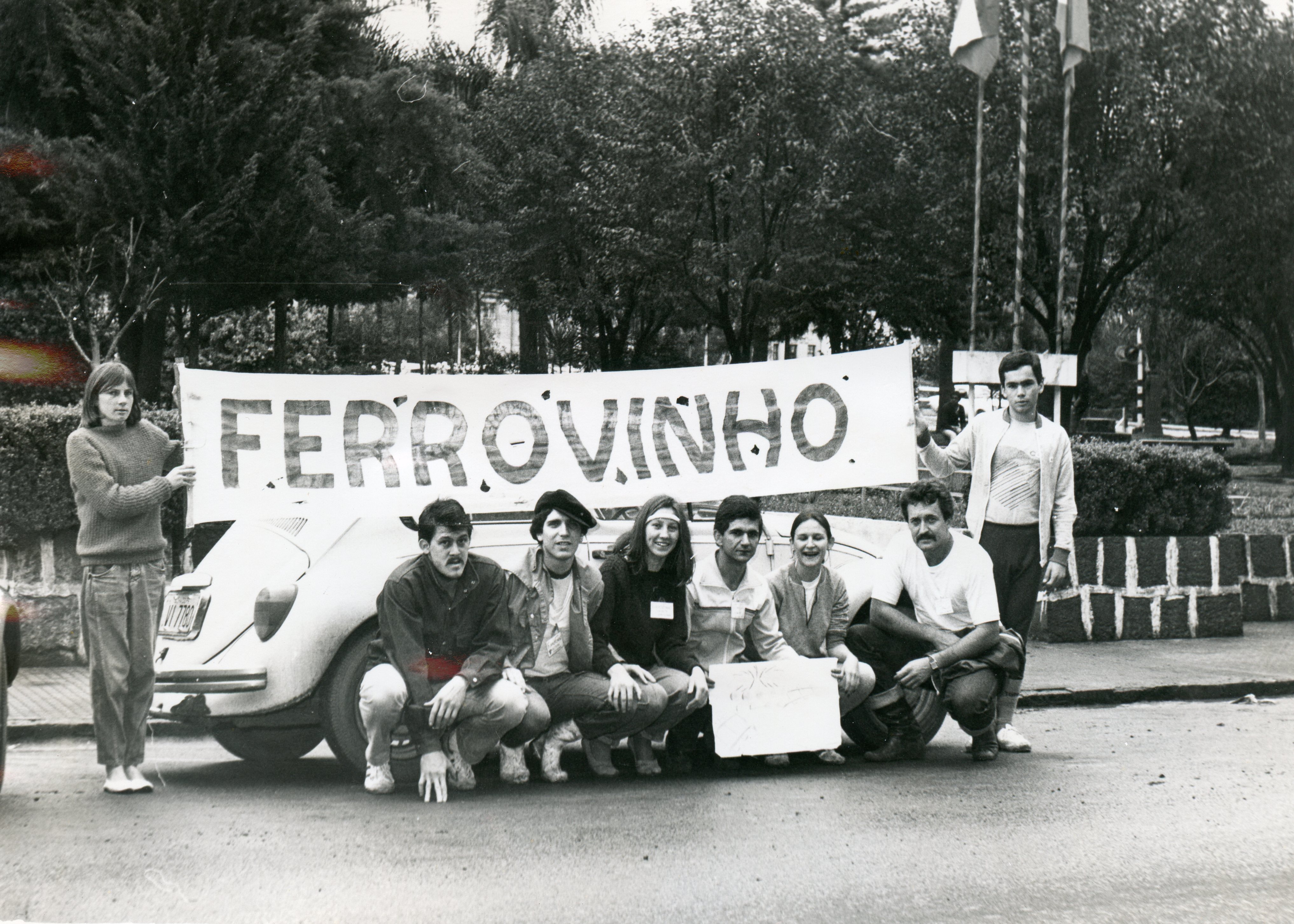 Uma fotografia em preto e branco mostrando um grupo de jovens posando atrás de um Volkswagen Fusca com uma faixa escrita 'FERROVINHO'. A cena parece ocorrer em uma praça ou área pública arborizada, sugerindo uma manifestação estudantil ou evento comunitário típico de uma cidade brasileira entre as décadas de 1970 e 1980.