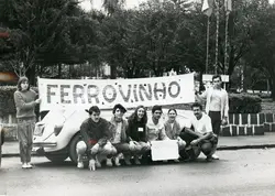 Uma fotografia em preto e branco mostrando um grupo de jovens posando atrás de um Volkswagen Fusca com uma faixa escrita 'FERROVINHO'. A cena parece ocorrer em uma praça ou área pública arborizada, sugerindo uma manifestação estudantil ou evento comunitário típico de uma cidade brasileira entre as décadas de 1970 e 1980.