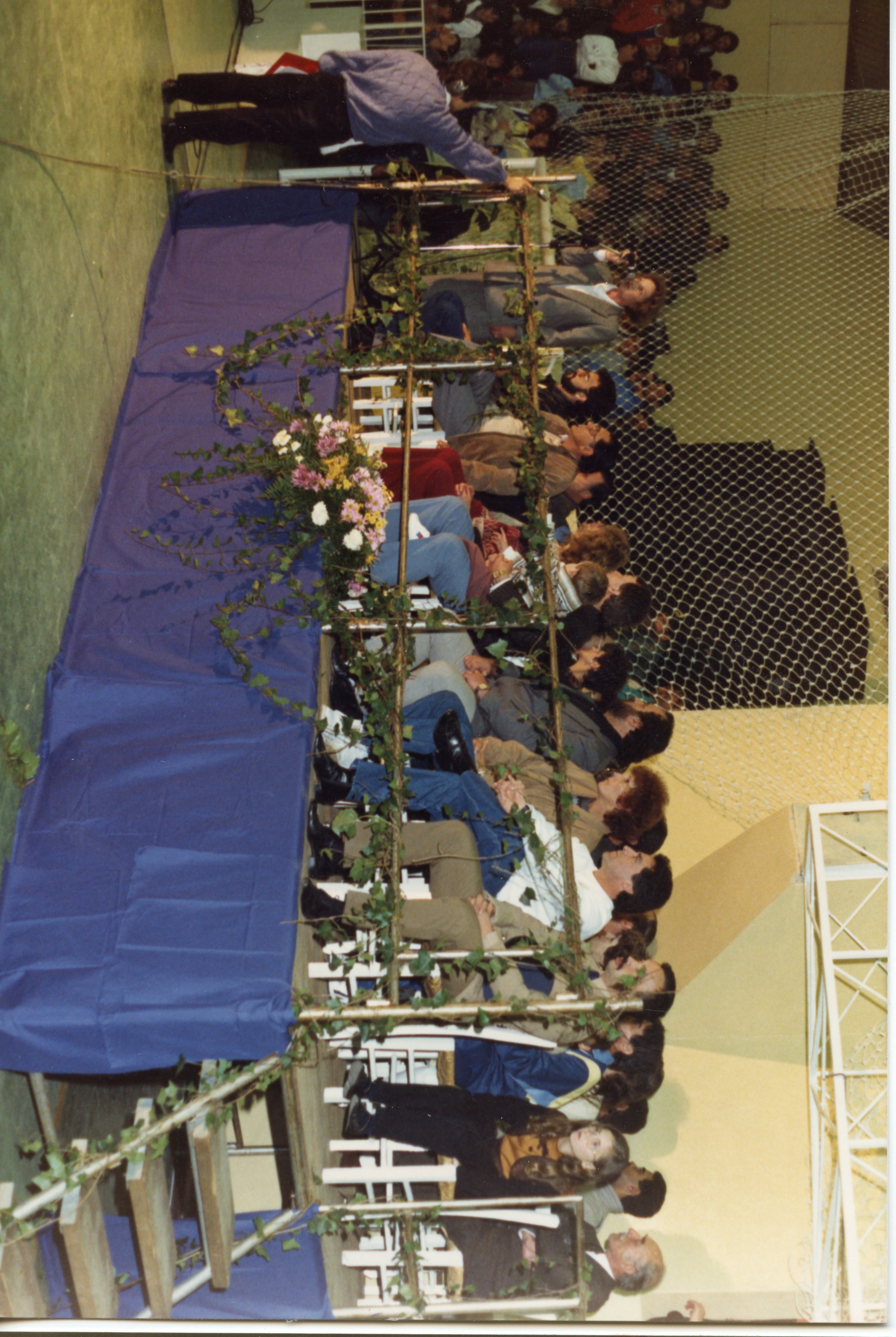 Uma fotografia capturada em um ângulo lateral mostrando um grupo de pessoas sentadas em uma mesa de honra ou palco decorado com flores e trepadeiras. O ambiente parece ser um ginásio ou salão comunitário, com uma rede de proteção ao fundo, sugerindo um evento social ou esportivo ocorrido provavelmente entre as décadas de 1980 e 1990.