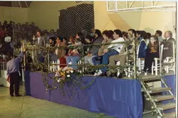 Uma fotografia capturada em um ângulo lateral mostrando um grupo de pessoas sentadas em uma mesa de honra ou palco decorado com flores e trepadeiras. O ambiente parece ser um ginásio ou salão comunitário, com uma rede de proteção ao fundo, sugerindo um evento social ou esportivo ocorrido provavelmente entre as décadas de 1980 e 1990.