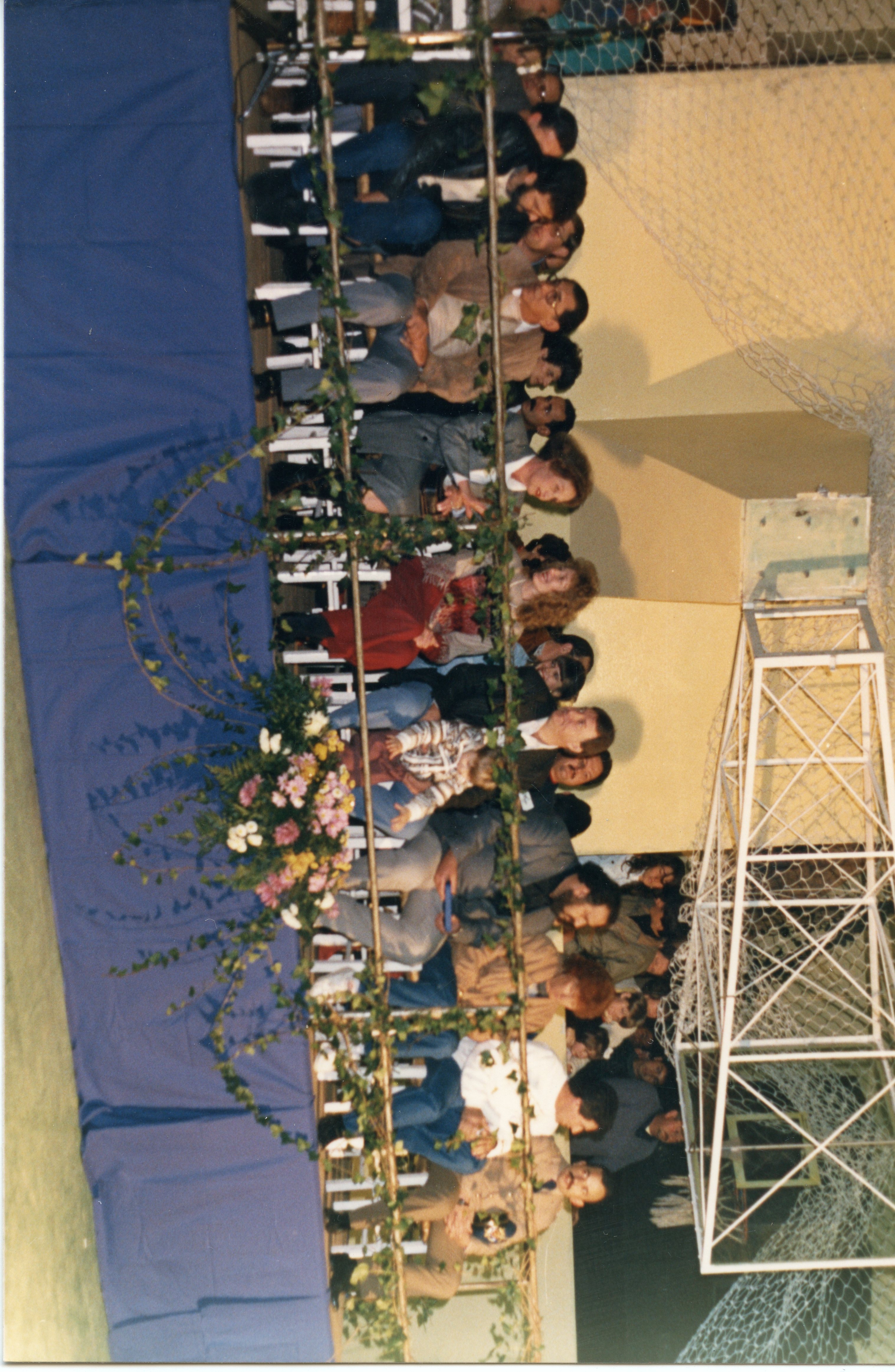 Uma fotografia que captura um grupo de pessoas sentadas em uma arquibancada ou área de plateia, possivelmente em um evento esportivo ou social em um ginásio. A imagem apresenta elementos decorativos como trepadeiras e flores, sugerindo uma ocasião especial, com vestimentas típicas das décadas de 1980 ou 1990.