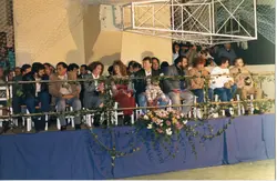 Uma fotografia que captura um grupo de pessoas sentadas em uma arquibancada ou área de plateia, possivelmente em um evento esportivo ou social em um ginásio. A imagem apresenta elementos decorativos como trepadeiras e flores, sugerindo uma ocasião especial, com vestimentas típicas das décadas de 1980 ou 1990.