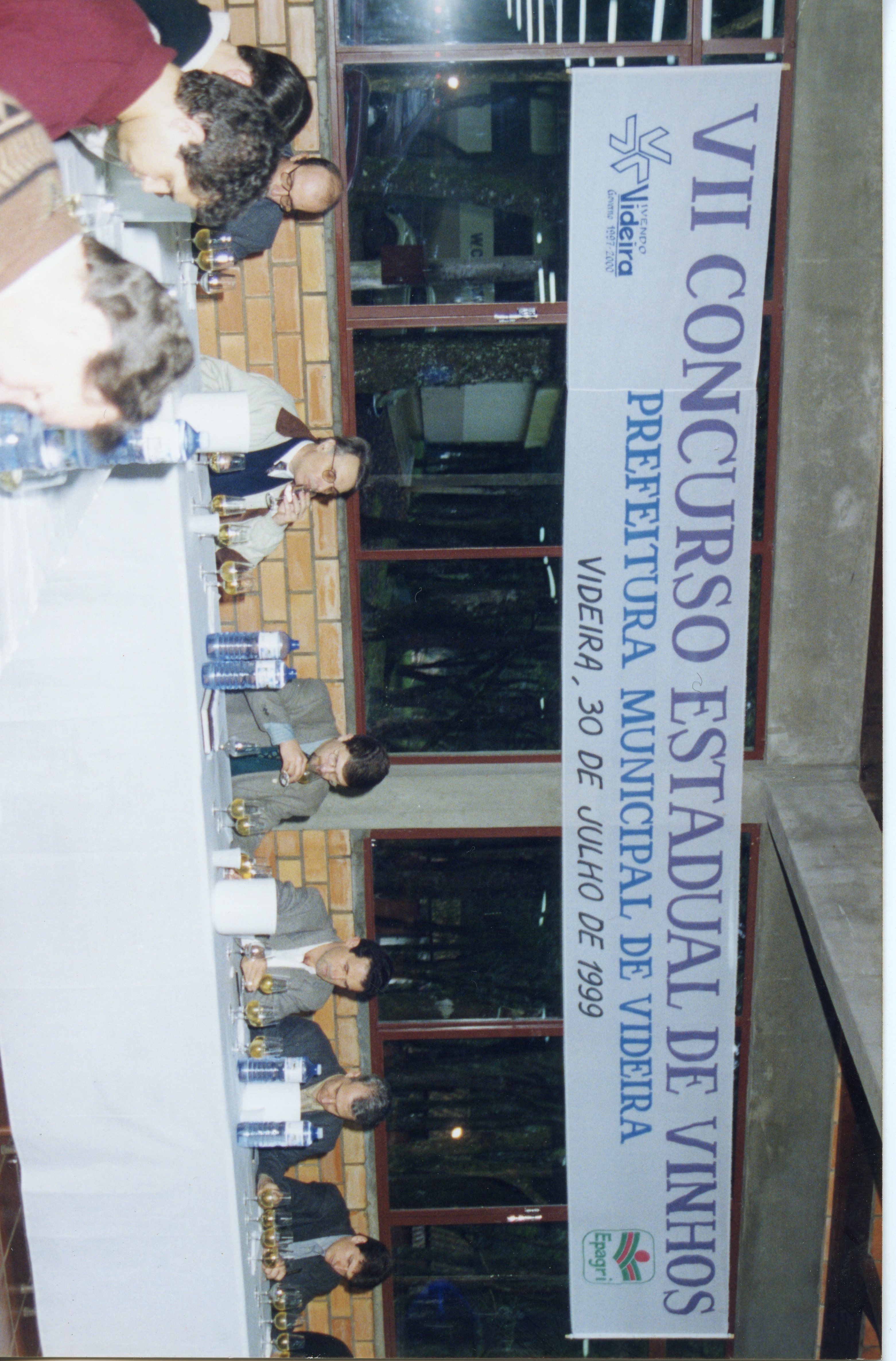 Fotografia de um painel ou banner vertical referente ao VII Concurso Estadual de Vinhos realizado em Videira, Santa Catarina. O evento, datado de 30 de julho de 1999, mostra um grupo de jurados ou participantes sentados a uma mesa longa realizando uma degustação técnica.