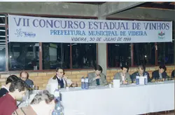 Fotografia de um painel ou banner vertical referente ao VII Concurso Estadual de Vinhos realizado em Videira, Santa Catarina. O evento, datado de 30 de julho de 1999, mostra um grupo de jurados ou participantes sentados a uma mesa longa realizando uma degustação técnica.