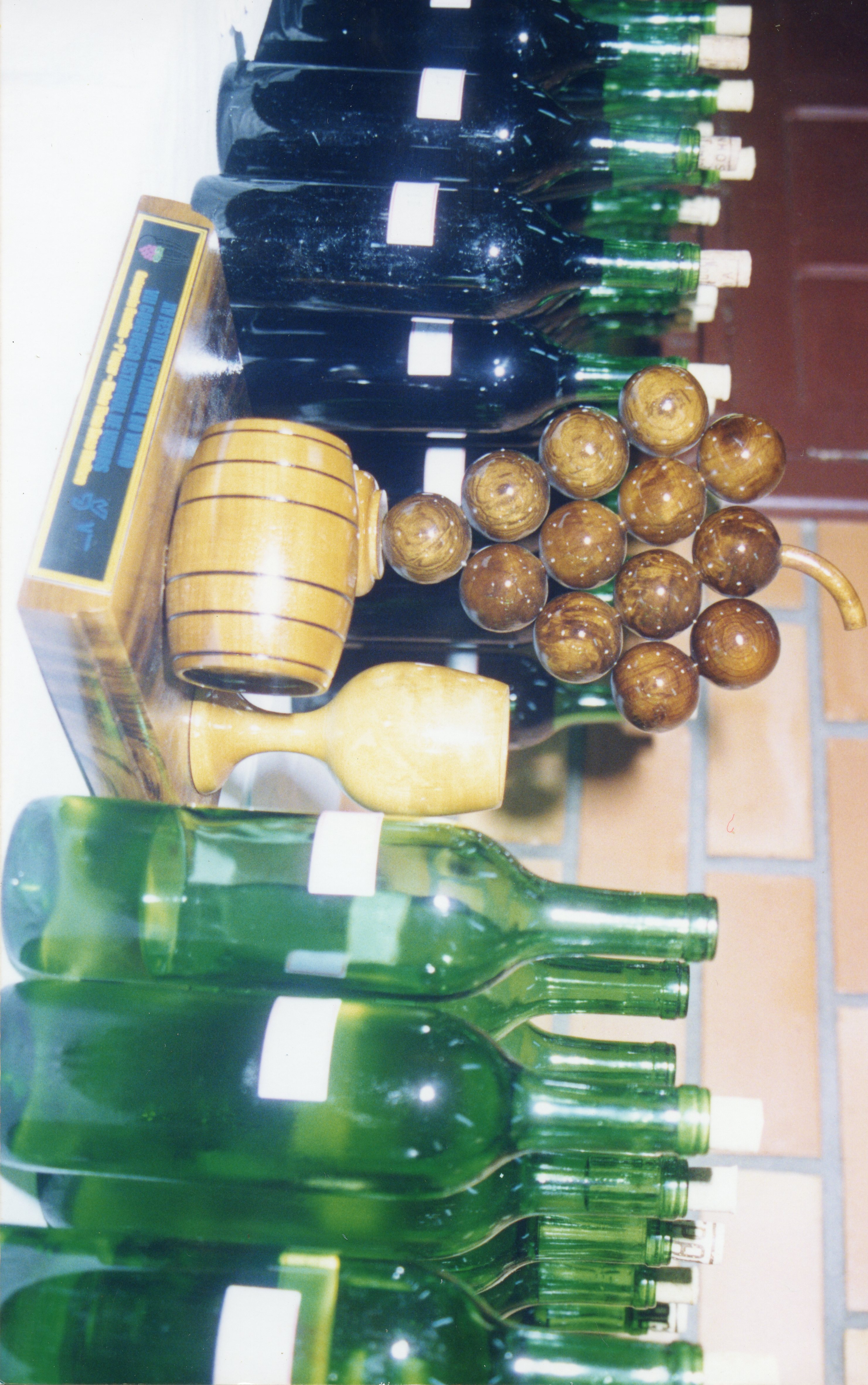 A imagem exibe uma coleção de garrafas de vinho alinhadas em uma prateleira, acompanhadas por uma peça decorativa de madeira em formato de barril e um cacho de uvas esculpido. O contexto sugere uma vinícola ou evento de celebração vitivinícola, possivelmente na região de Videira, conhecida por sua tradição na produção de vinhos.