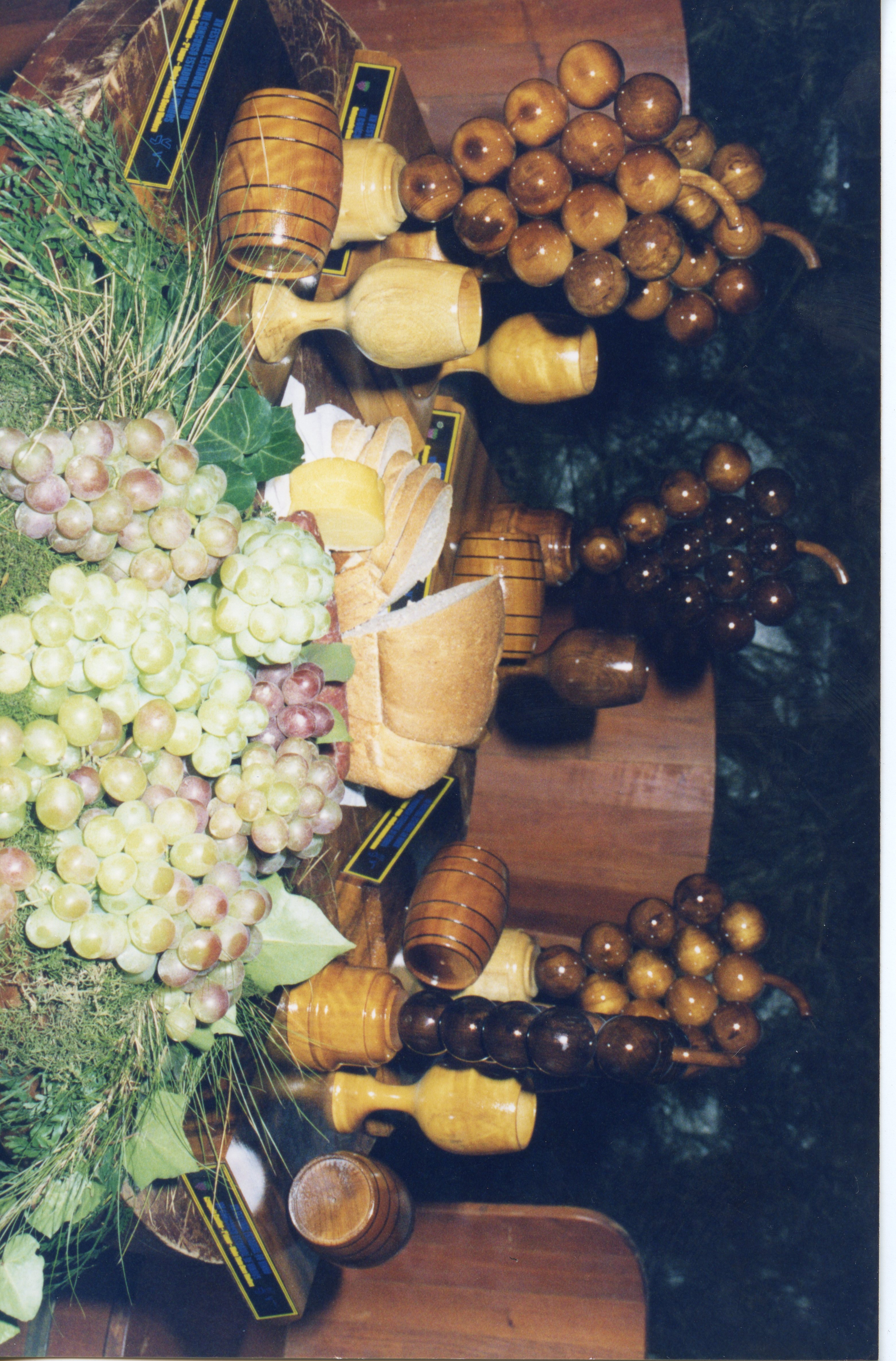 Uma composição de mesa exibindo elementos típicos da cultura vitivinícola, incluindo cachos de uvas frescas, pães fatiados e réplicas artesanais de madeira em formato de uvas, barris e taças. A cena sugere um contexto de exposição cultural ou gastronômica, possivelmente relacionada a festividades de colheita em uma região como Videira.