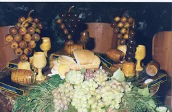 Uma composição de mesa exibindo elementos típicos da cultura vitivinícola, incluindo cachos de uvas frescas, pães fatiados e réplicas artesanais de madeira em formato de uvas, barris e taças. A cena sugere um contexto de exposição cultural ou gastronômica, possivelmente relacionada a festividades de colheita em uma região como Videira.