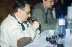 Uma fotografia que captura um momento de degustação técnica de vinhos, possivelmente em um evento profissional ou concurso. Homens de meia-idade estão sentados à mesa com taças de vinho tinto e papéis de avaliação, sugerindo um contexto de análise enológica comum em regiões vitivinícolas como Videira.