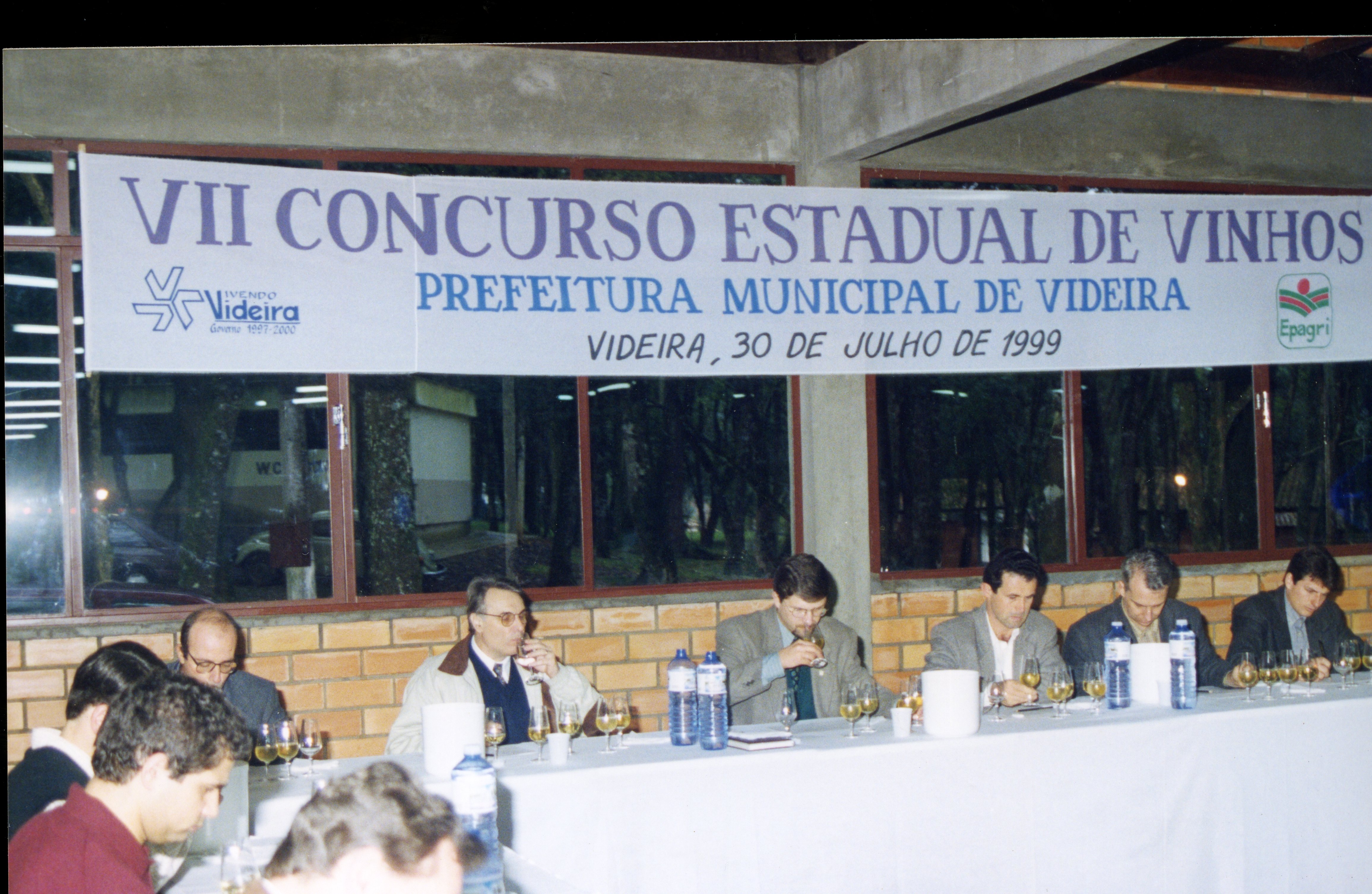 Uma fotografia que registra o VII Concurso Estadual de Vinhos realizado em Videira, Santa Catarina, em 1999. A imagem mostra um grupo de jurados sentados a uma mesa longa, degustando vinhos, com uma faixa oficial da Prefeitura Municipal ao fundo.