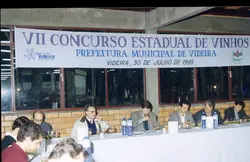 Uma fotografia que registra o VII Concurso Estadual de Vinhos realizado em Videira, Santa Catarina, em 1999. A imagem mostra um grupo de jurados sentados a uma mesa longa, degustando vinhos, com uma faixa oficial da Prefeitura Municipal ao fundo.
