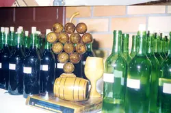 Uma fotografia colorida exibindo um troféu de madeira em formato de cacho de uvas e barril, posicionado sobre uma mesa entre diversas garrafas de vinho. O troféu apresenta uma placa comemorativa, sugerindo um evento de premiação vitivinícola, possivelmente relacionado à cultura da uva e do vinho em uma região como Videira.