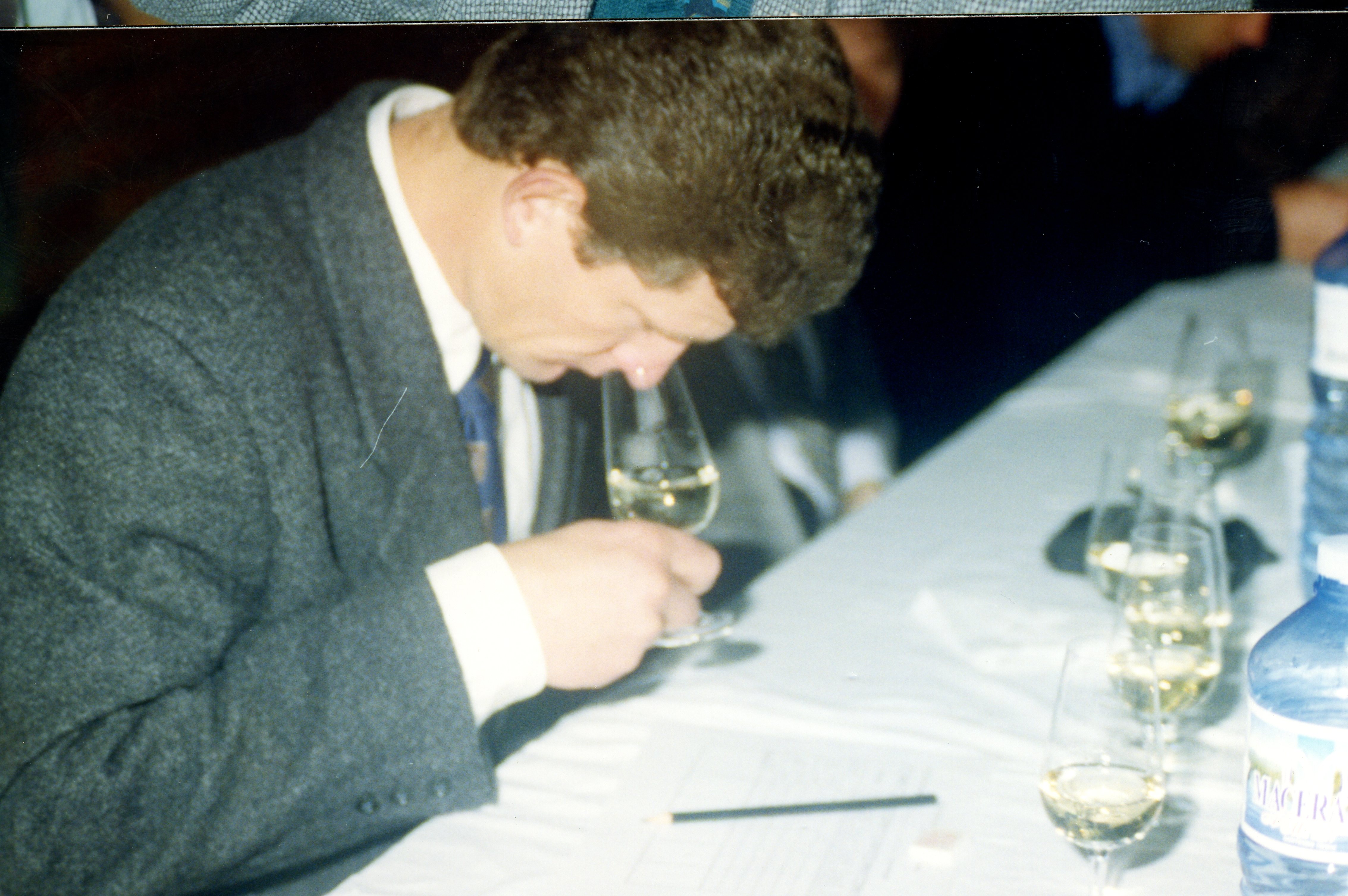 Uma fotografia que captura um homem de terno cinza concentrado em uma degustação de vinhos, segurando uma taça para avaliar o aroma. O ambiente parece ser um evento formal ou técnico, com várias taças alinhadas sobre uma mesa coberta por uma toalha branca, sugerindo uma análise profissional ou concurso de vinhos.