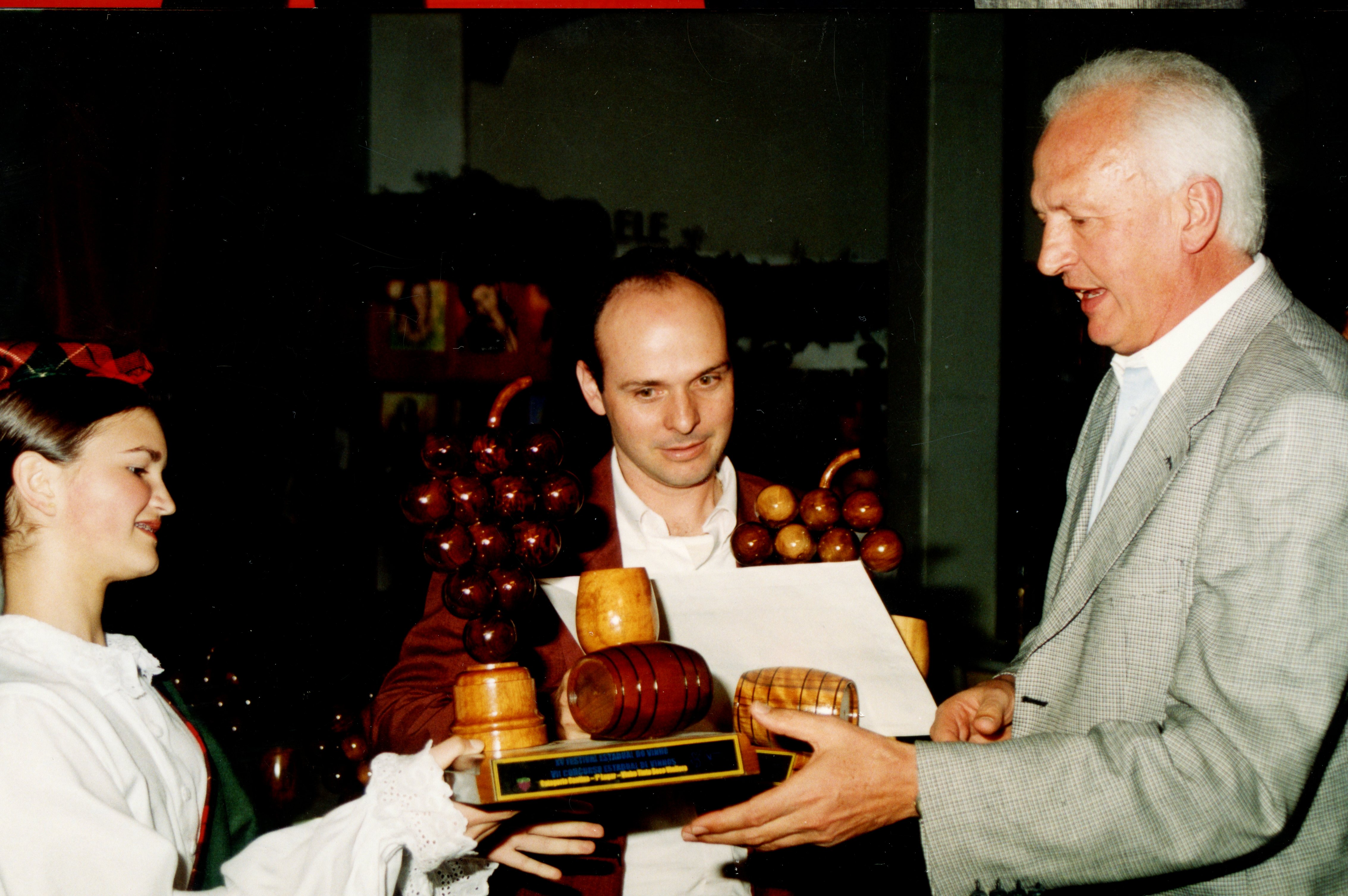 Uma fotografia que registra a entrega de uma premiação, possivelmente relacionada a um festival de uvas ou vitivinicultura, dado o formato dos troféus em madeira. A cena mostra três pessoas em um ambiente interno, com vestimentas formais e tradicionais, sugerindo um evento comemorativo regional, possivelmente em uma cidade com forte influência da imigração italiana como Videira.