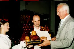 Uma fotografia que registra a entrega de uma premiação, possivelmente relacionada a um festival de uvas ou vitivinicultura, dado o formato dos troféus em madeira. A cena mostra três pessoas em um ambiente interno, com vestimentas formais e tradicionais, sugerindo um evento comemorativo regional, possivelmente em uma cidade com forte influência da imigração italiana como Videira.