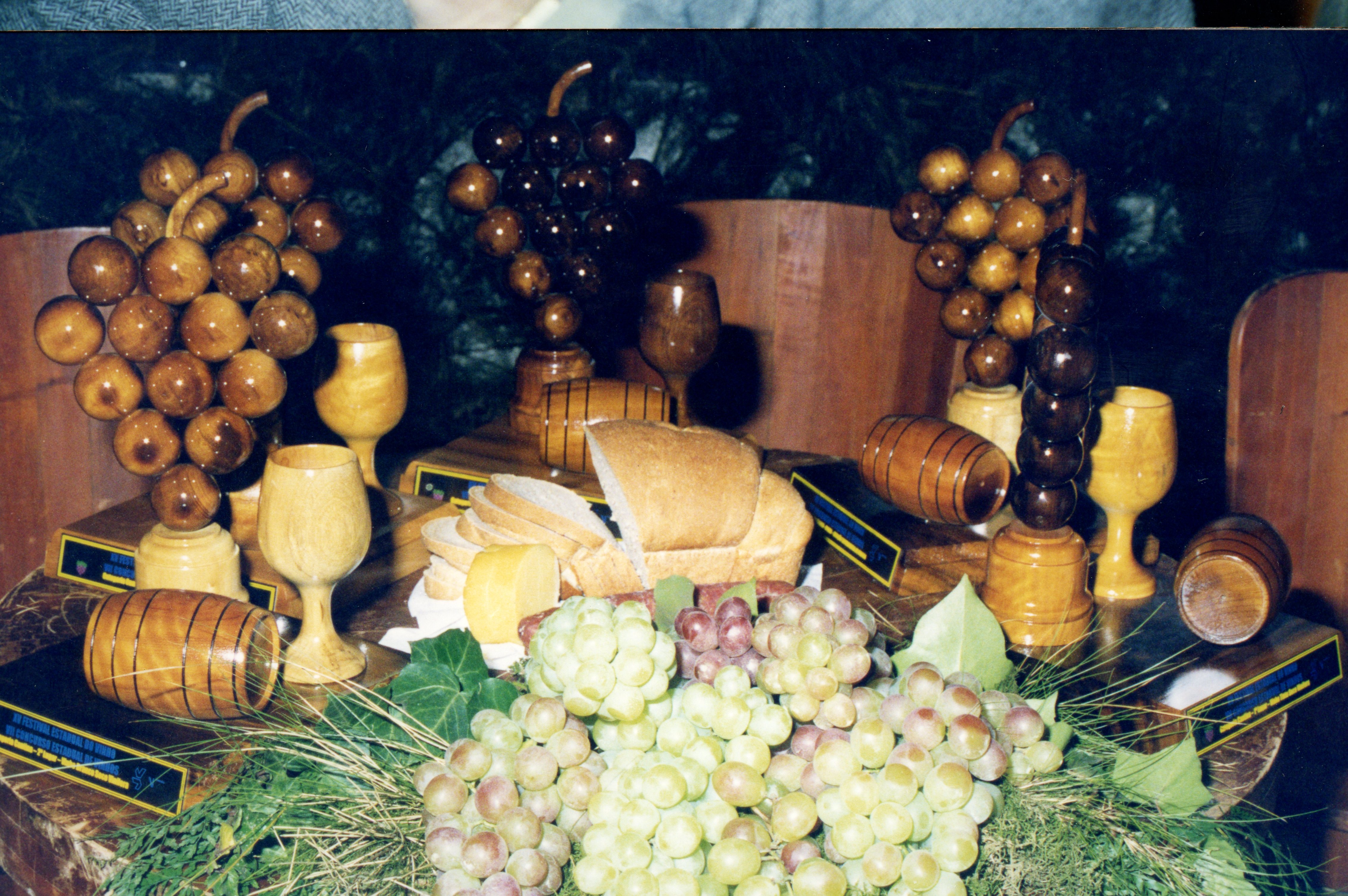 Uma mesa decorada com elementos que remetem à vitivinicultura, incluindo esculturas de madeira em formato de cachos de uva, taças e pequenos barris. A cena sugere um contexto de celebração ou festival de uvas, possivelmente em uma região como Videira, destacando a cultura local e a produção artesanal.