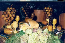 Uma mesa decorada com elementos que remetem à vitivinicultura, incluindo esculturas de madeira em formato de cachos de uva, taças e pequenos barris. A cena sugere um contexto de celebração ou festival de uvas, possivelmente em uma região como Videira, destacando a cultura local e a produção artesanal.