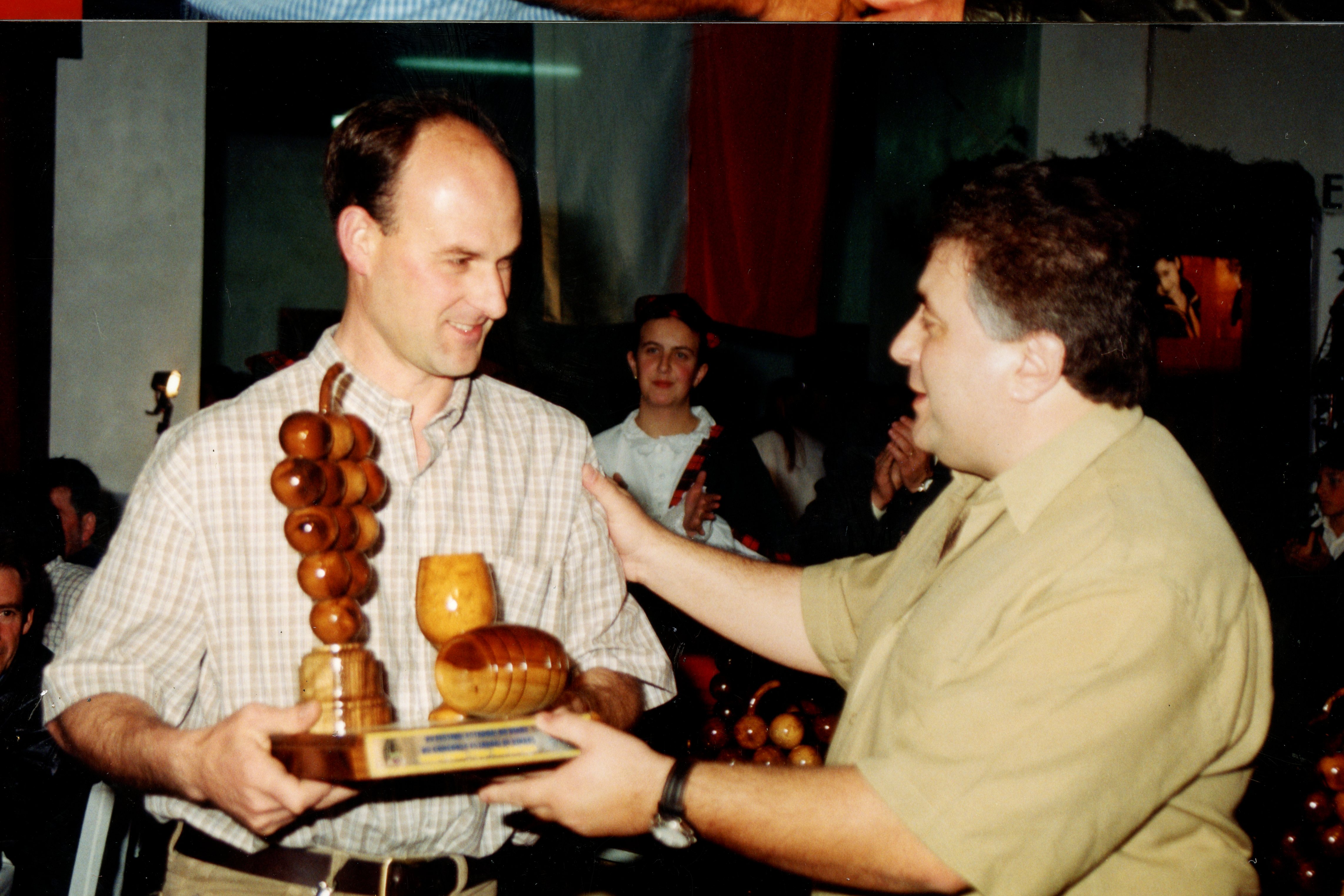 Uma fotografia que registra a entrega de um troféu de madeira em formato de uvas e barril, sugerindo um evento relacionado à vitivinicultura, possivelmente em Videira. A imagem mostra dois homens em um ambiente interno, com uma mulher ao fundo aplaudindo, indicando uma cerimônia de premiação ou homenagem.