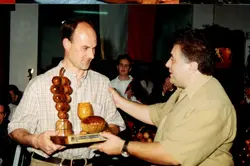 Uma fotografia que registra a entrega de um troféu de madeira em formato de uvas e barril, sugerindo um evento relacionado à vitivinicultura, possivelmente em Videira. A imagem mostra dois homens em um ambiente interno, com uma mulher ao fundo aplaudindo, indicando uma cerimônia de premiação ou homenagem.