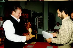 Uma fotografia que registra um momento de entrega de premiação ou homenagem, onde um homem sorridente recebe um troféu de madeira em formato de uvas. O cenário sugere um evento social ou cultural, possivelmente relacionado à viticultura, comum em regiões como Videira, com vestimentas típicas do final do século XX.