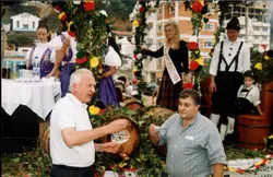 Uma cena festiva ao ar livre que parece ser um desfile ou celebração cultural, possivelmente uma Festa do Vinho, dado o traje típico e a faixa da rainha. O evento apresenta pessoas vestidas com roupas tradicionais germânicas, barris de madeira decorados com flores e folhagens, e um ambiente urbano ao fundo, sugerindo uma celebração comunitária em uma cidade com forte influência da imigração europeia.