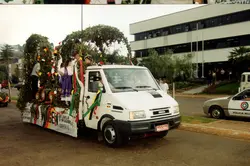 Uma fotografia colorida de um desfile comemorativo, provavelmente em Videira, Santa Catarina, destacando um caminhão Iveco decorado com elementos temáticos de uvas e folhagens. Pessoas vestidas com trajes típicos estão sobre a carroceria, sugerindo uma celebração da cultura da imigração italiana e da vitivinicultura local, possivelmente durante a Festa da Uva.
