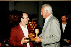 Uma fotografia que registra a entrega de um troféu de madeira em formato de cacho de uvas e barril, simbolizando a vitivinicultura. A cena sugere um evento formal ou premiação, possivelmente relacionado à cultura da uva em uma região como Videira, com dois homens em destaque e um terceiro ao fundo.