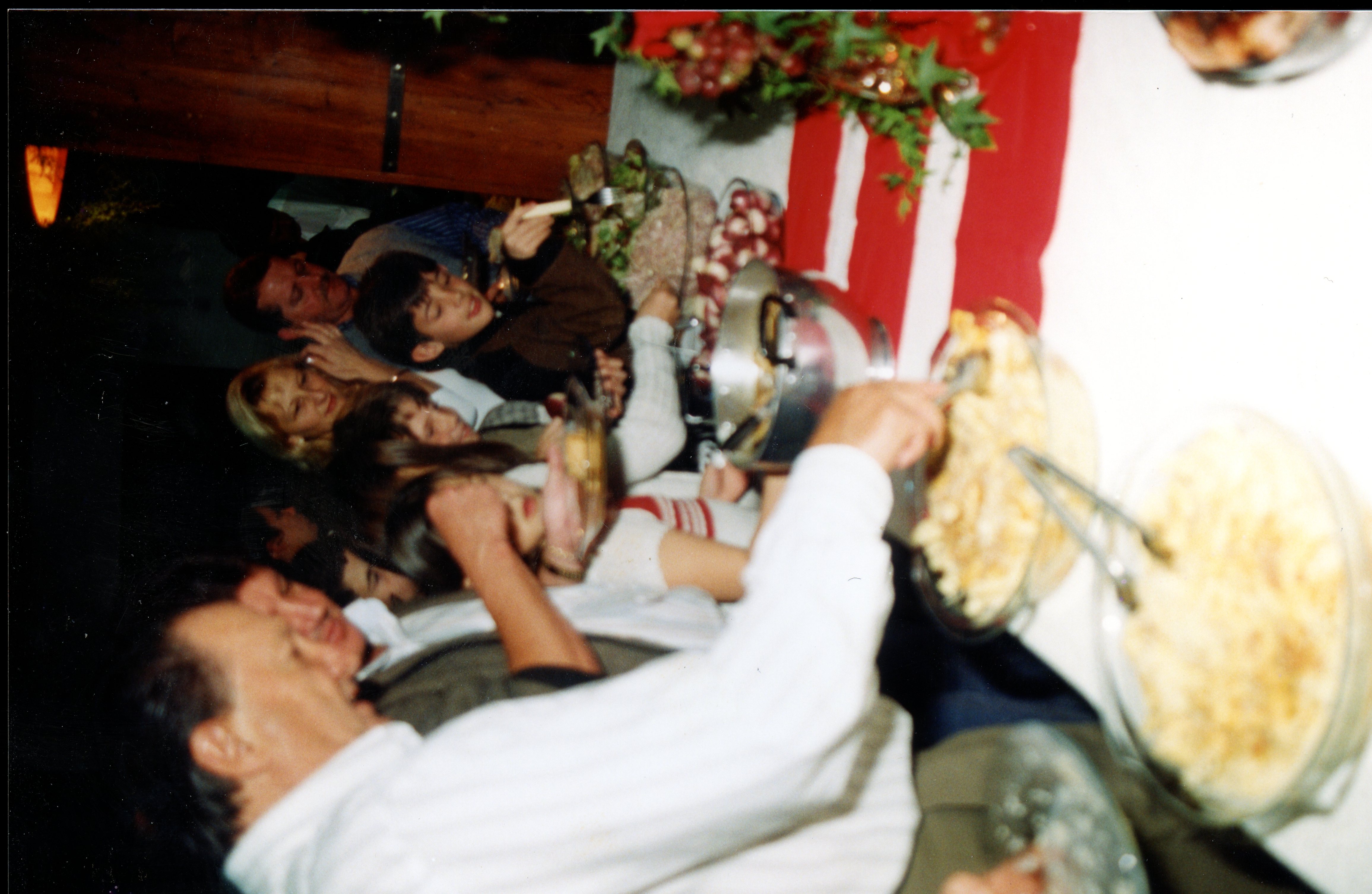 Uma fotografia de um jantar ou confraternização familiar em um ambiente interno, possivelmente nas décadas de 1980 ou 1990. Várias pessoas estão sentadas ao redor de uma mesa farta, servindo-se de pratos típicos, sugerindo um evento social ou comemorativo em um contexto doméstico ou comunitário.