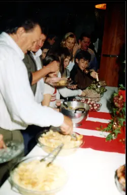 Uma fotografia de um jantar ou confraternização familiar em um ambiente interno, possivelmente nas décadas de 1980 ou 1990. Várias pessoas estão sentadas ao redor de uma mesa farta, servindo-se de pratos típicos, sugerindo um evento social ou comemorativo em um contexto doméstico ou comunitário.
