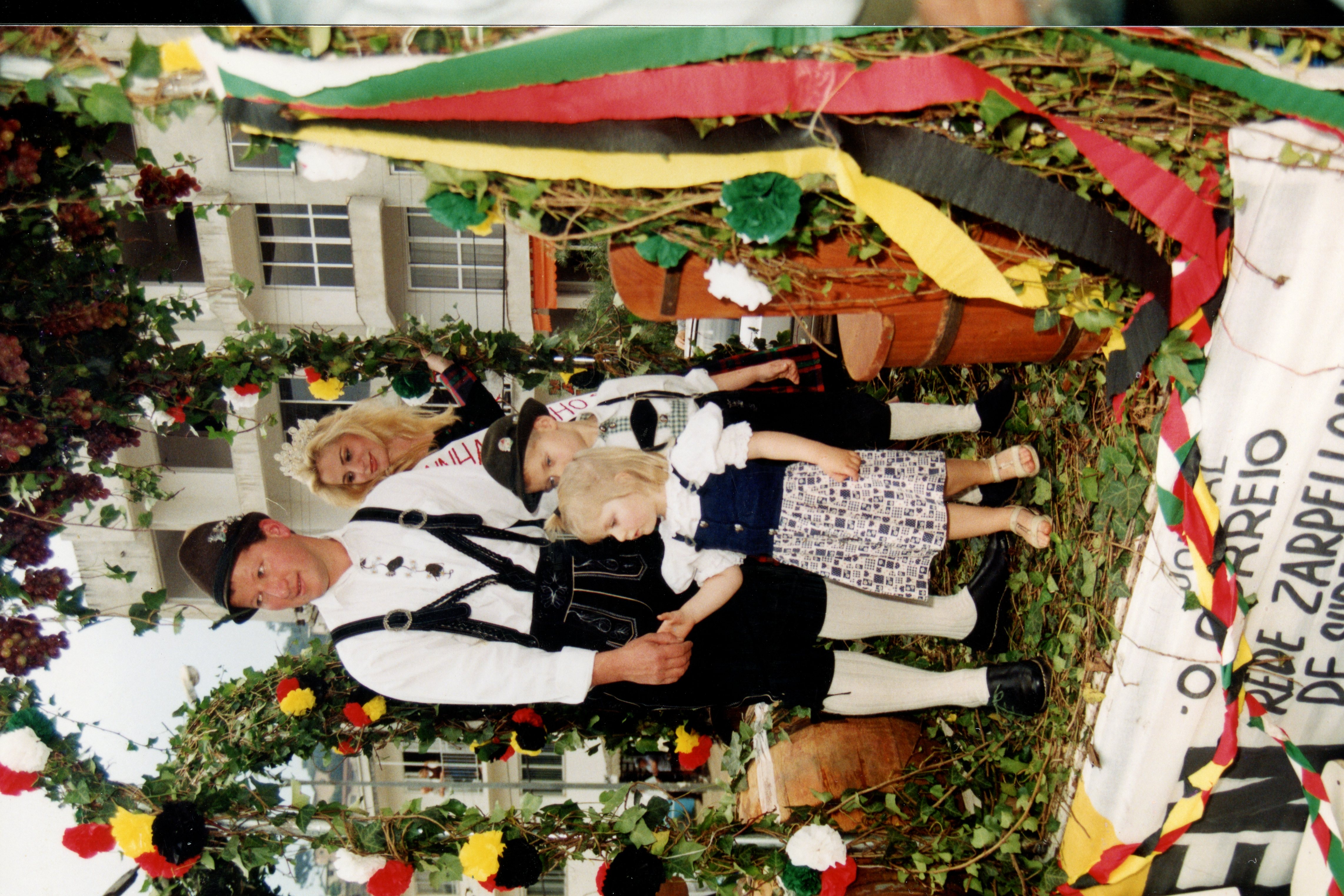 Uma fotografia colorida que retrata uma família vestida com trajes típicos alemães, possivelmente participando de um desfile cultural ou festa da uva em uma cidade como Videira. O cenário inclui uma estrutura decorada com folhagens e uvas, sugerindo um evento de celebração da imigração europeia, com elementos arquitetônicos urbanos ao fundo.