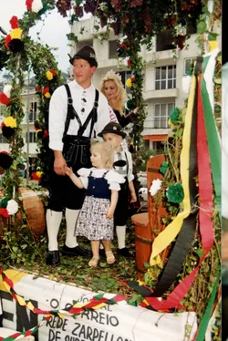 Uma fotografia colorida que retrata uma família vestida com trajes típicos alemães, possivelmente participando de um desfile cultural ou festa da uva em uma cidade como Videira. O cenário inclui uma estrutura decorada com folhagens e uvas, sugerindo um evento de celebração da imigração europeia, com elementos arquitetônicos urbanos ao fundo.