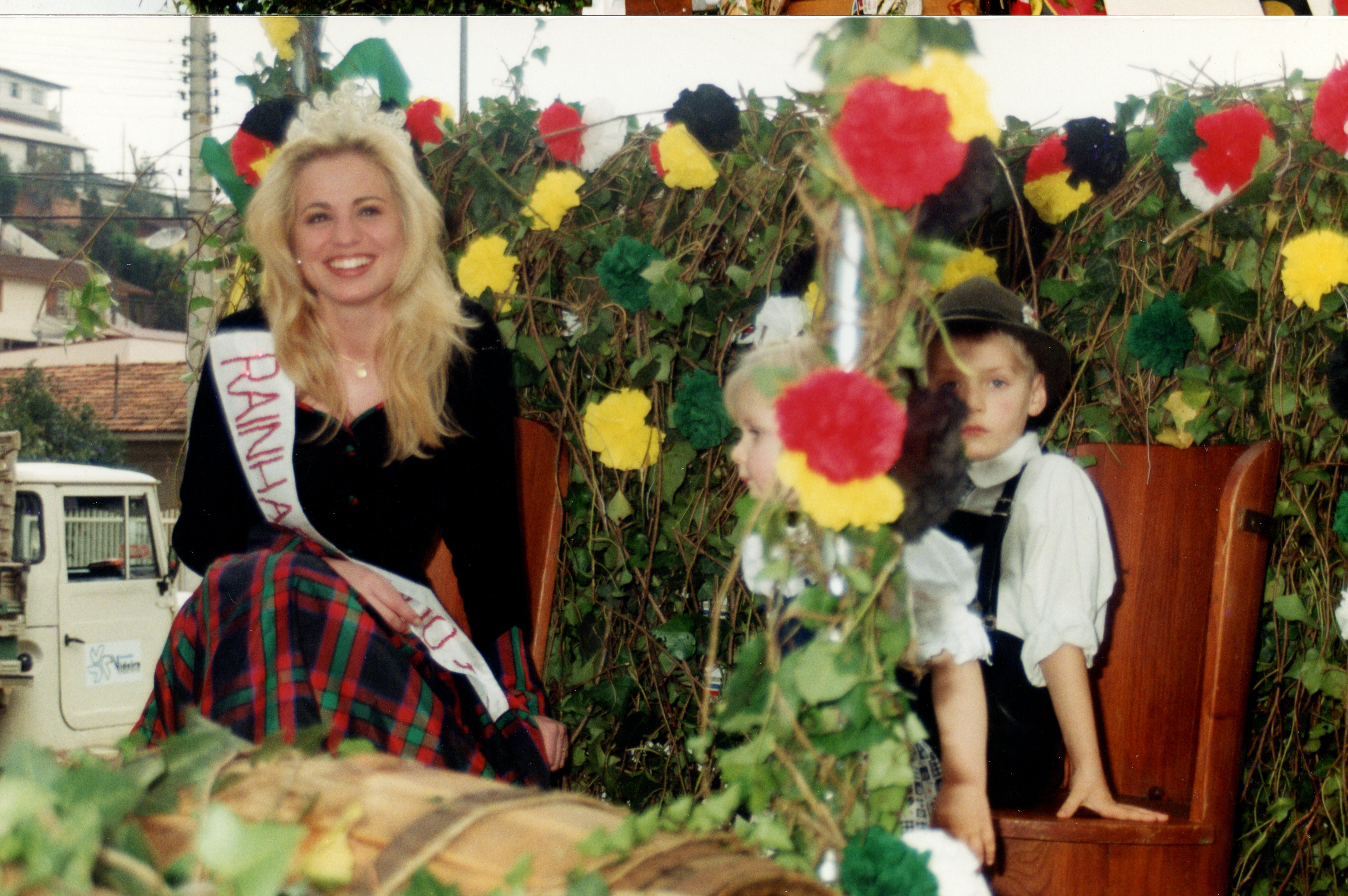 Uma fotografia colorida que retrata uma jovem mulher usando uma faixa de 'Rainha' e trajes típicos, sentada em um carro alegórico decorado com flores e folhagens. Ao seu lado, duas crianças vestidas com trajes tradicionais germânicos, sugerindo um evento cultural ou desfile típico em uma cidade com forte influência da imigração europeia, possivelmente no sul do Brasil.