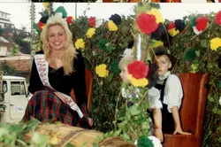 Uma fotografia colorida que retrata uma jovem mulher usando uma faixa de 'Rainha' e trajes típicos, sentada em um carro alegórico decorado com flores e folhagens. Ao seu lado, duas crianças vestidas com trajes tradicionais germânicos, sugerindo um evento cultural ou desfile típico em uma cidade com forte influência da imigração europeia, possivelmente no sul do Brasil.