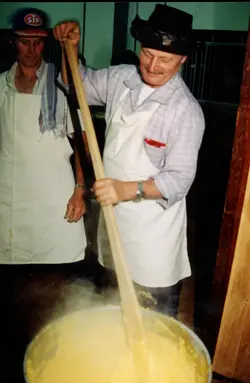 Dois homens vestindo aventais brancos preparam uma grande quantidade de polenta em um caldeirão, utilizando uma pá de madeira. A cena sugere um evento comunitário ou gastronômico tradicional, possivelmente em uma região de colonização italiana no Brasil, como Videira, durante a década de 1980 ou 1990.