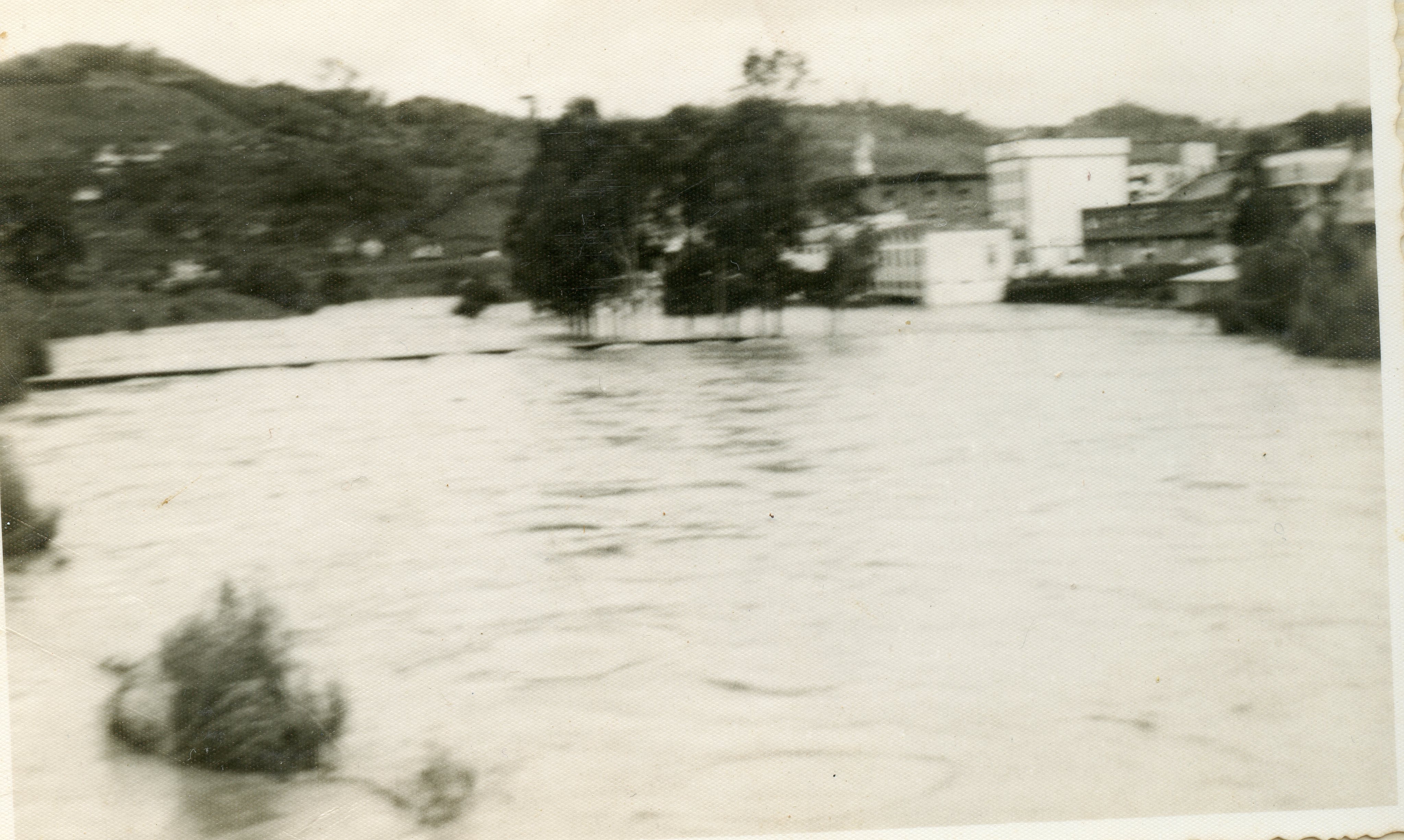 Fotografia em preto e branco registrando uma enchente em uma área urbana, possivelmente em uma cidade do interior como Videira. A imagem mostra um rio transbordado cobrindo ruas e vegetação, com edificações ao fundo parcialmente cercadas pela água.
