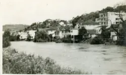 Fotografia em preto e branco que retrata uma paisagem urbana ou suburbana situada às margens de um rio ou curso d'água. A imagem exibe construções residenciais simples em uma encosta, sugerindo um cenário de desenvolvimento urbano inicial ou uma pequena cidade brasileira, possivelmente da metade do século XX.