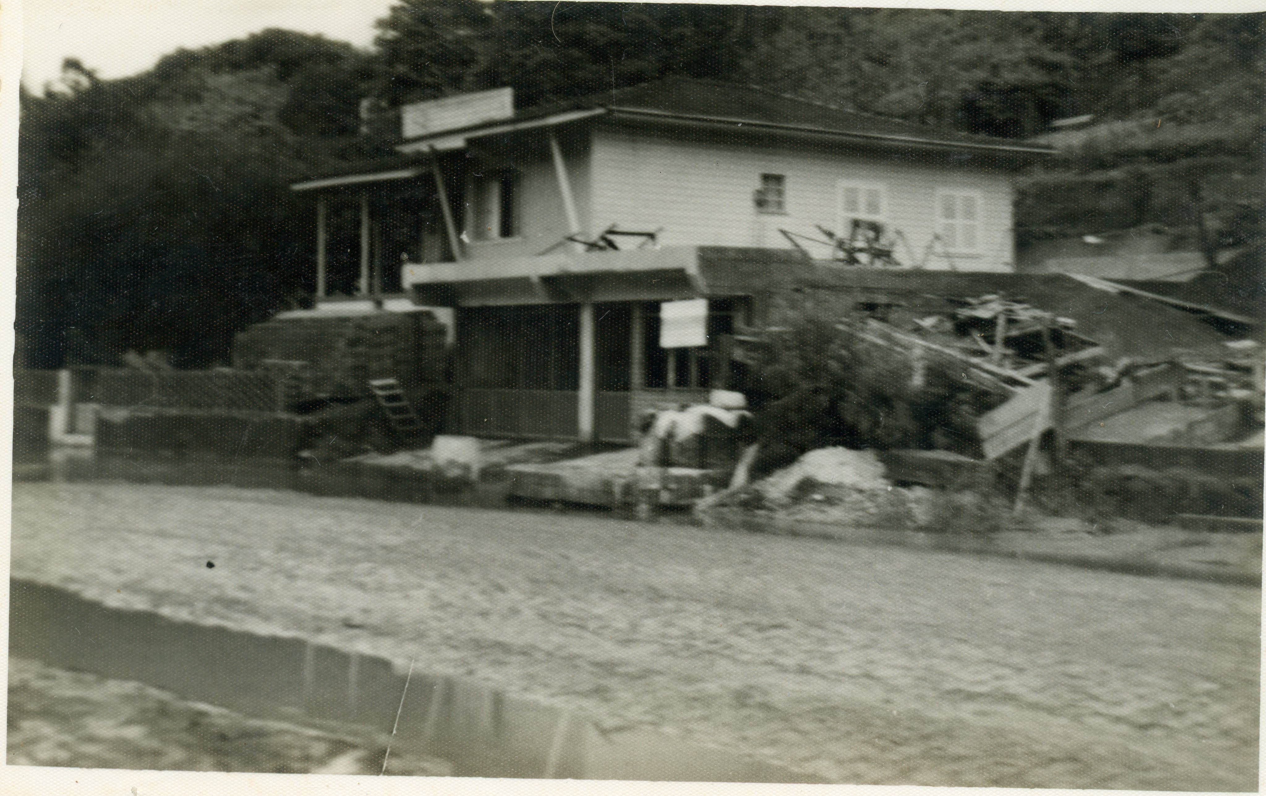Fotografia em preto e branco de uma construção de madeira com base em alvenaria, possivelmente um estabelecimento comercial ou residência em uma área rural ou pequena cidade brasileira. A imagem apresenta sinais de desgaste e parece capturar um momento de construção ou reforma, com materiais espalhados ao redor da estrutura.