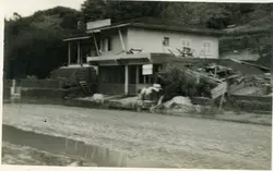 Fotografia em preto e branco de uma construção de madeira com base em alvenaria, possivelmente um estabelecimento comercial ou residência em uma área rural ou pequena cidade brasileira. A imagem apresenta sinais de desgaste e parece capturar um momento de construção ou reforma, com materiais espalhados ao redor da estrutura.