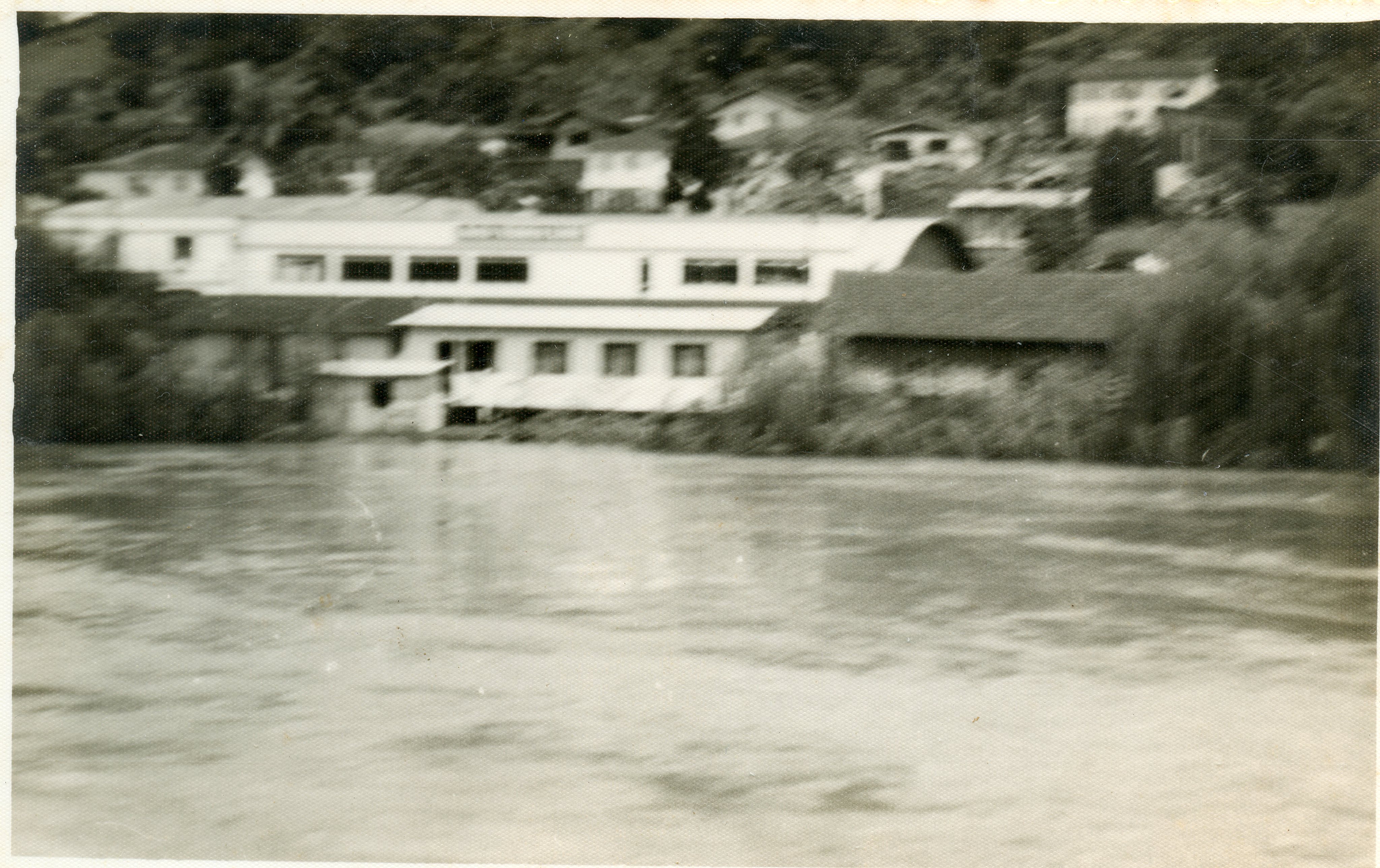 Fotografia antiga em preto e branco mostrando uma construção industrial ou comercial situada às margens de um rio. A imagem apresenta um aspecto rural ou de pequena cidade, com casas dispersas na encosta ao fundo e uma estrutura de telhado curvo característica de construções de meados do século XX.
