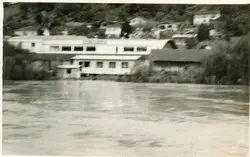 Fotografia antiga em preto e branco mostrando uma construção industrial ou comercial situada às margens de um rio. A imagem apresenta um aspecto rural ou de pequena cidade, com casas dispersas na encosta ao fundo e uma estrutura de telhado curvo característica de construções de meados do século XX.