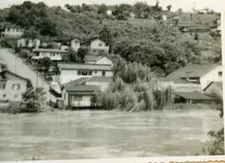 Fotografia em preto e branco mostrando uma área residencial situada em uma encosta, possivelmente em uma cidade do interior do Brasil como Videira. A imagem exibe casas de arquitetura simples dispostas em terreno inclinado, com vegetação densa e um corpo d'água ou área plana em primeiro plano, sugerindo um registro histórico de meados do século XX.