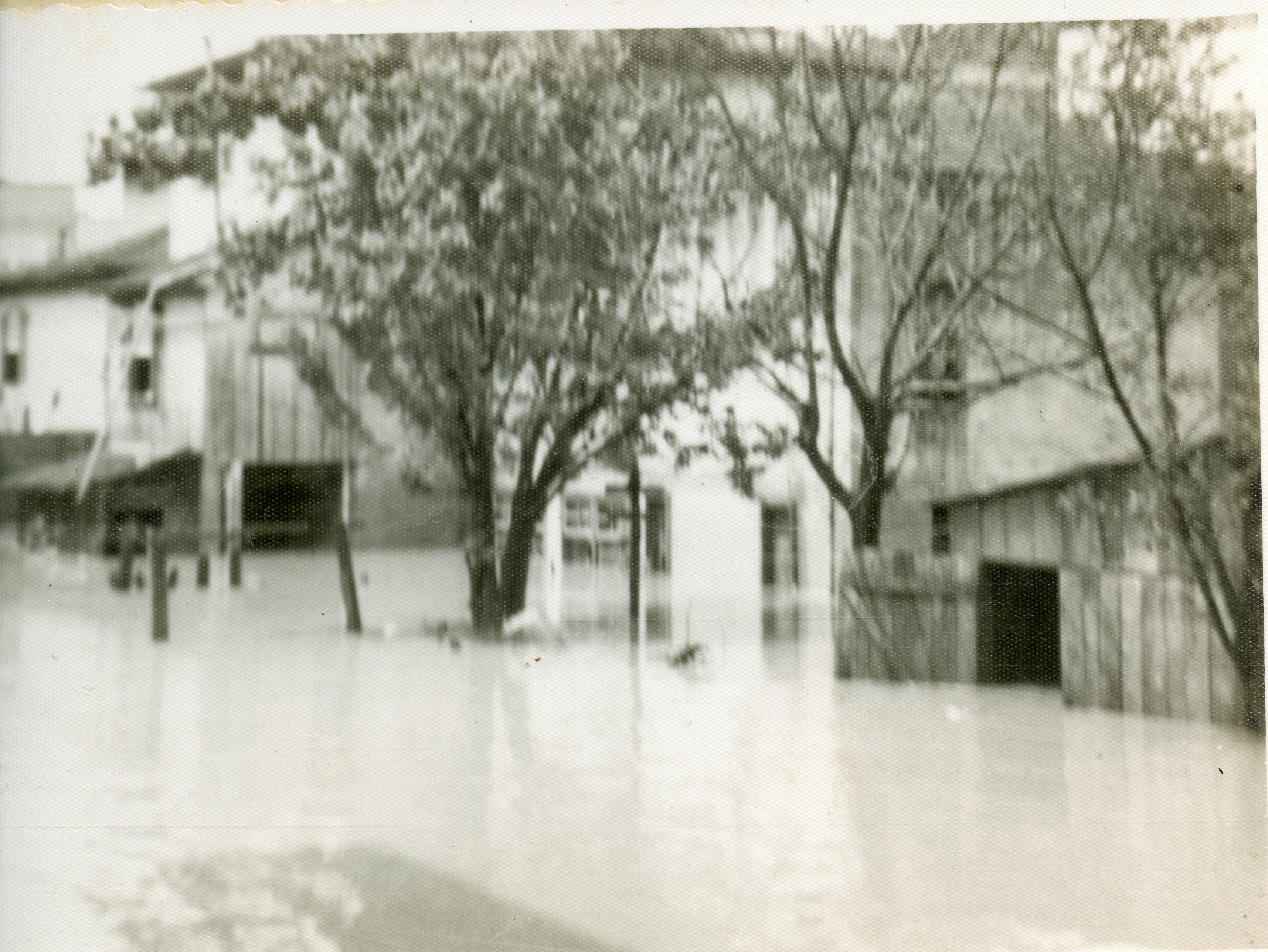 Fotografia em preto e branco que registra uma enchente em uma área urbana, possivelmente em uma pequena cidade brasileira. A imagem mostra casas parcialmente submersas e árvores cercadas pela água, sugerindo um evento climático severo ocorrido provavelmente em meados do século XX.