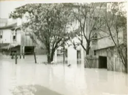 Fotografia em preto e branco que registra uma enchente em uma área urbana, possivelmente em uma pequena cidade brasileira. A imagem mostra casas parcialmente submersas e árvores cercadas pela água, sugerindo um evento climático severo ocorrido provavelmente em meados do século XX.