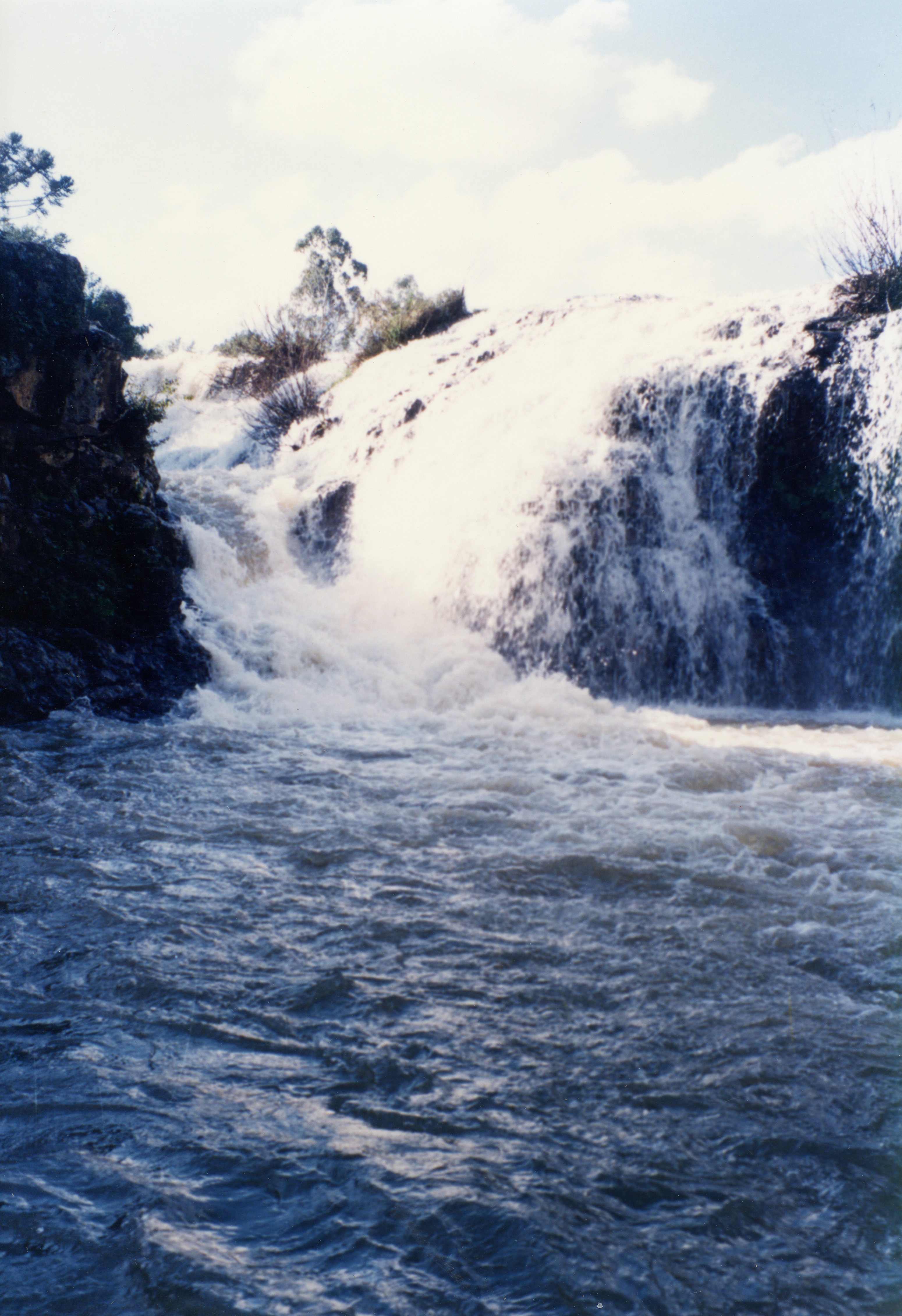 Esta imagem colorida retrata uma cachoeira com grande volume de água caindo sobre rochas e vegetação, formando um rio ou lago na parte inferior. O céu azul com algumas nuvens brancas é visível ao fundo. A qualidade da fotografia e as cores sugerem que pode ser uma imagem do final do século XX, possivelmente de uma área natural em uma região como Videira ou uma pequena cidade brasileira.